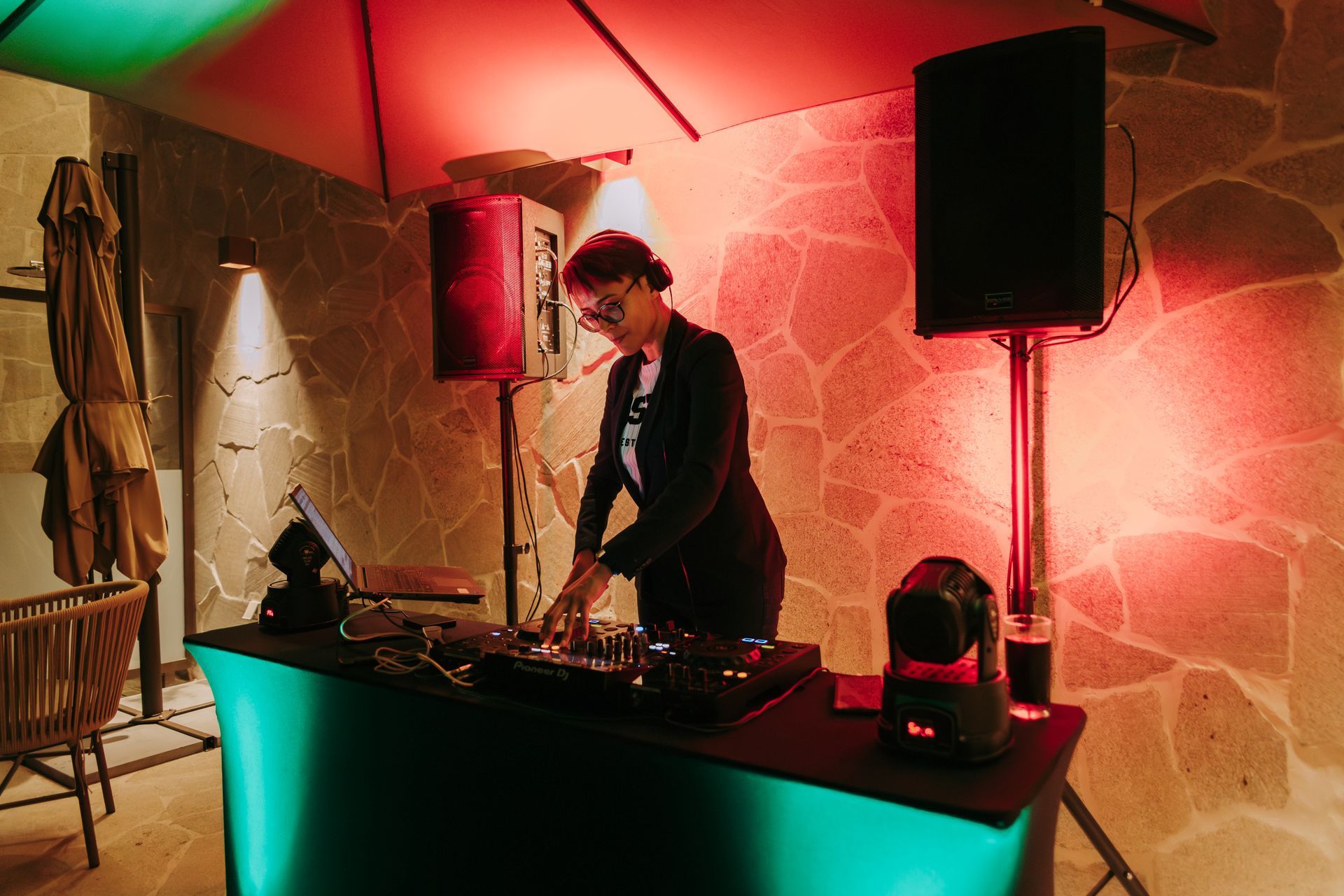 DJ em uma festa, operando equipamentos atrás de uma mesa preta com alto-falantes em uma sala bem iluminada. Othon