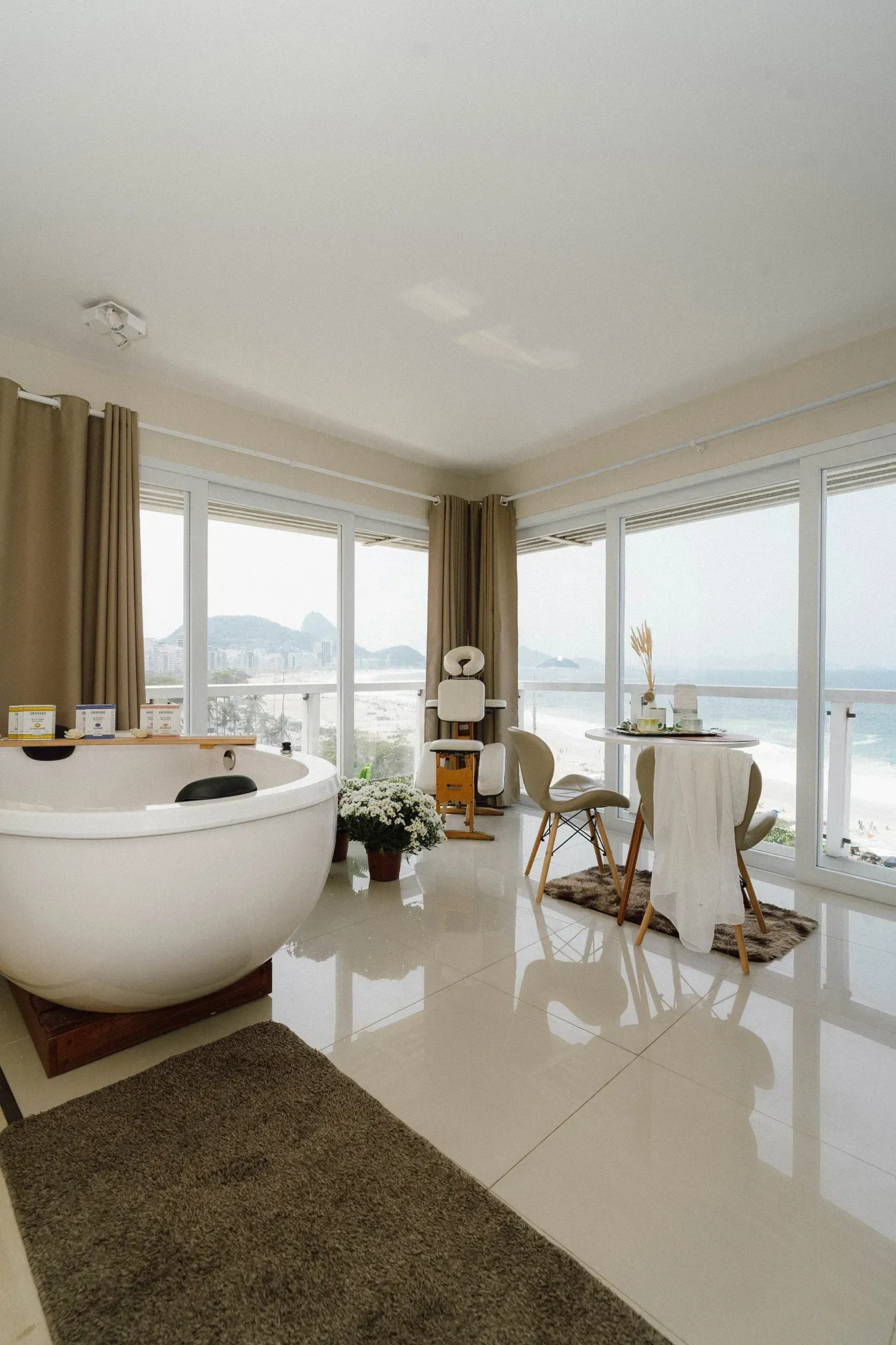 Banheiro estilo spa com banheira grande branca, tapete marrom, janelas do chão ao teto e vista para o mar.