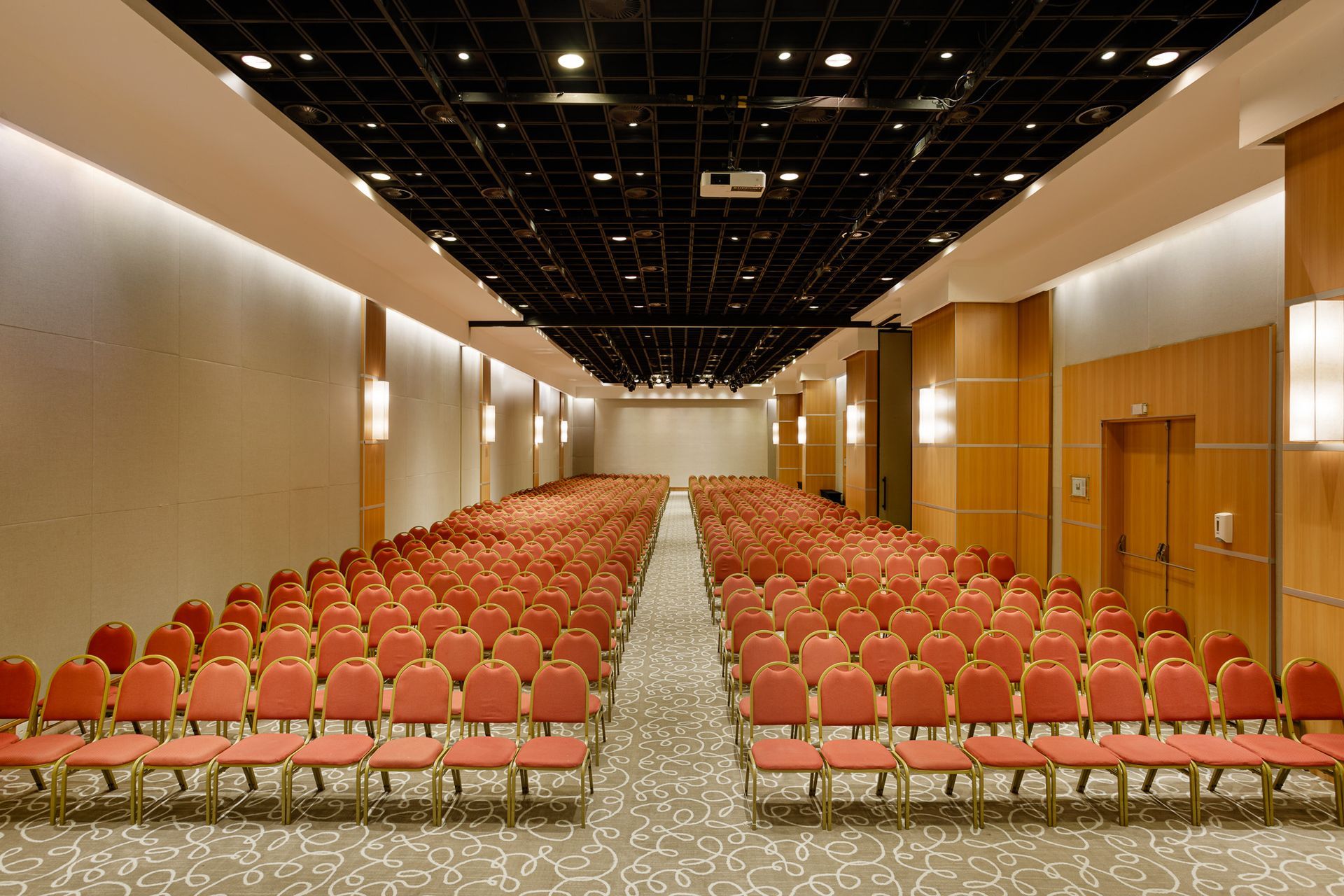 Auditório com fileiras de cadeiras vermelhas, paredes bege, teto escuro e tela de projeção.