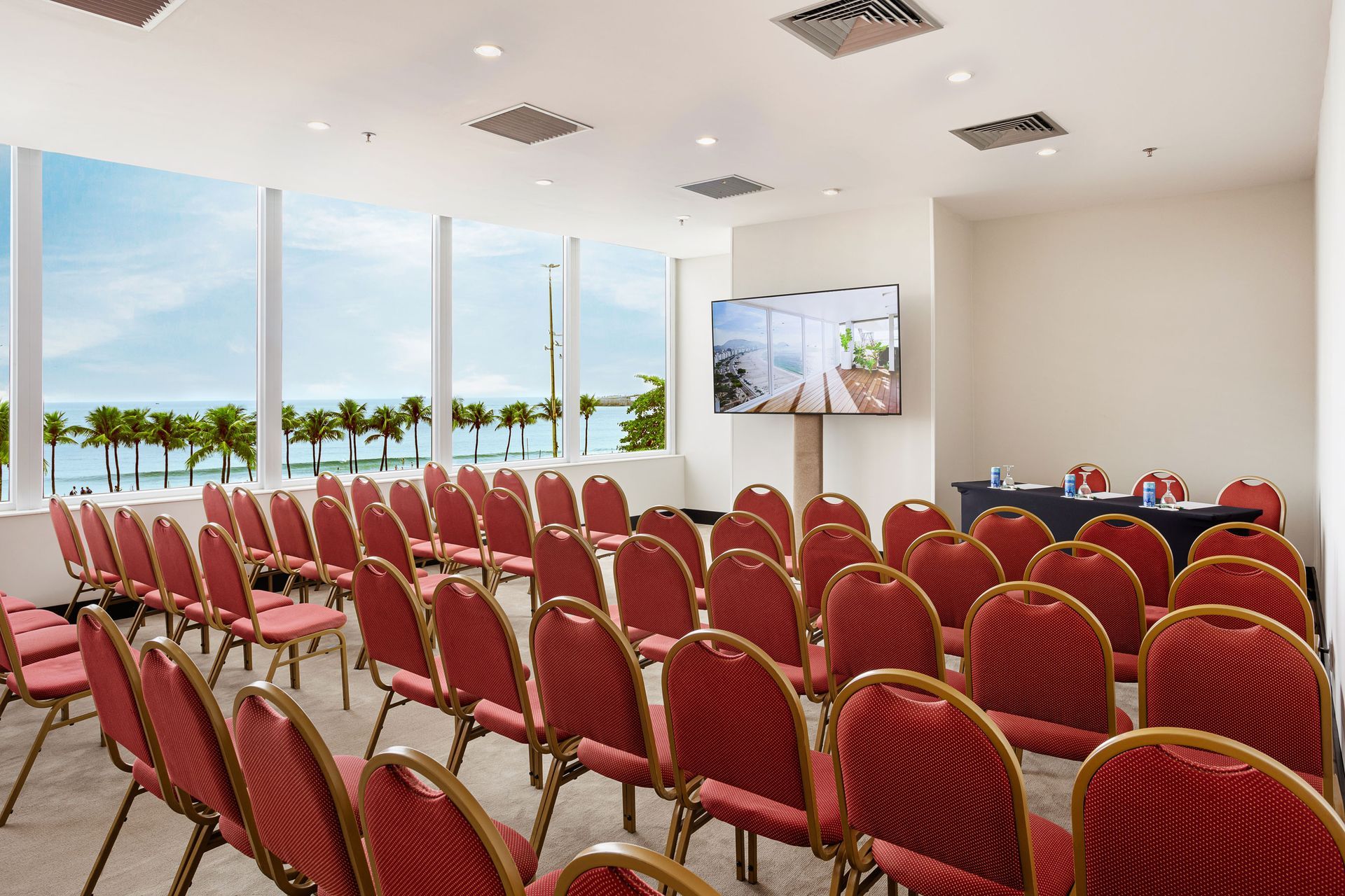 Fileiras de cadeiras vermelhas ficam de frente para uma mesa em uma sala de conferências com uma grande janela com vista para a praia. Othon