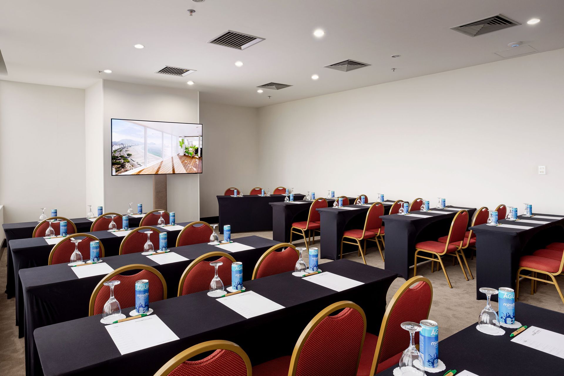 Sala de conferências com mesas, cadeiras e um telão, preparada para uma apresentação. Toalhas de mesa pretas, cadeiras vermelhas. Othon