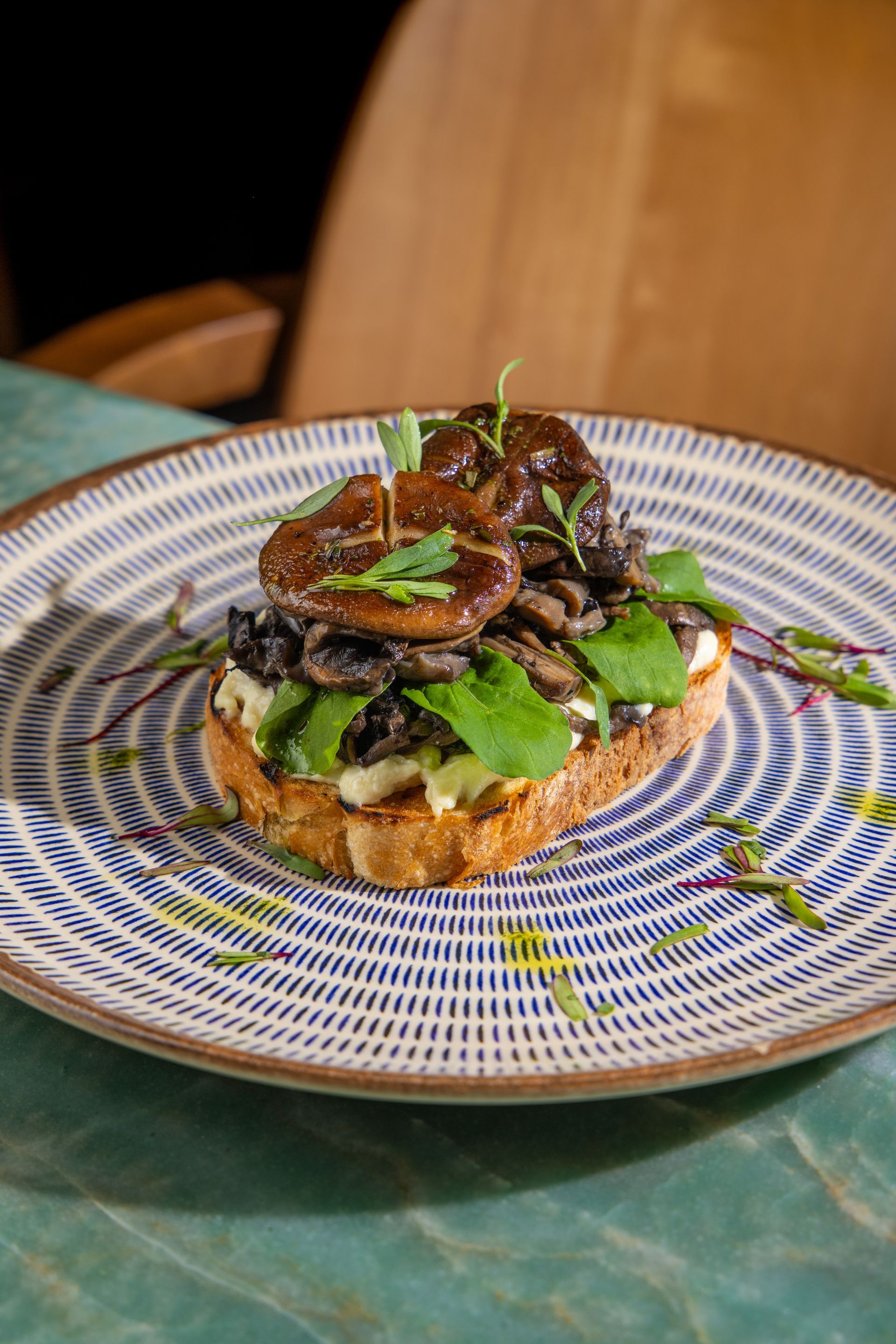 Torrada aberta com cogumelos servida em um prato estampado, coberta com folhas verdes, sobre um fundo azul-petróleo e madeira.