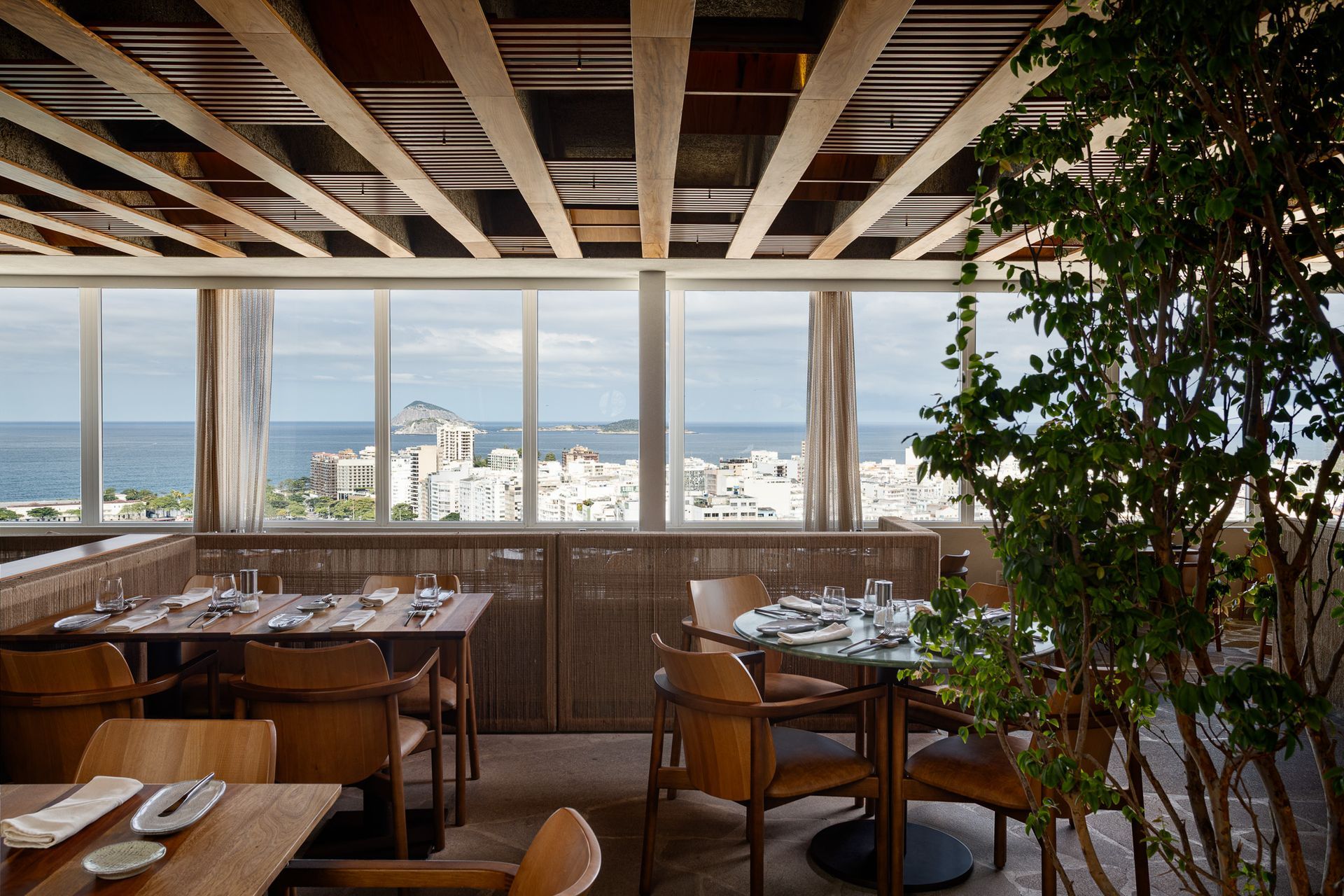 Interior de restaurante com mesas e cadeiras de madeira, vista da janela para o oceano e edifícios.