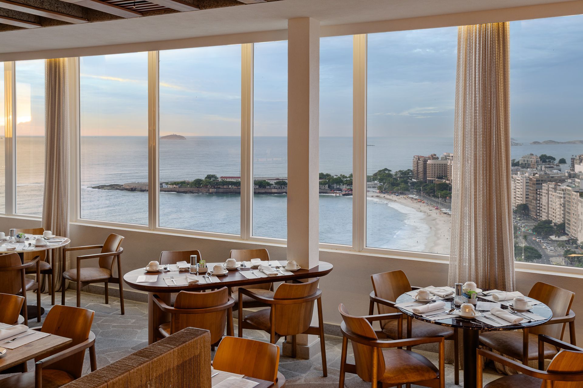 Interior de restaurante com mesas e cadeiras perto de grandes janelas com vista para o oceano e a praia.