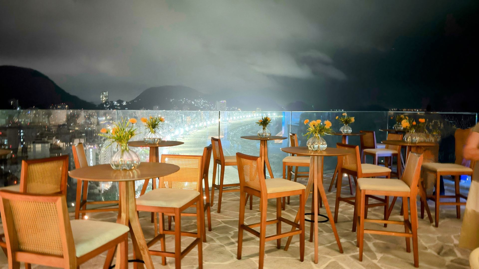 Bar na cobertura à noite com vista para a cidade. Mesas e cadeiras de madeira com arranjos florais. Othon