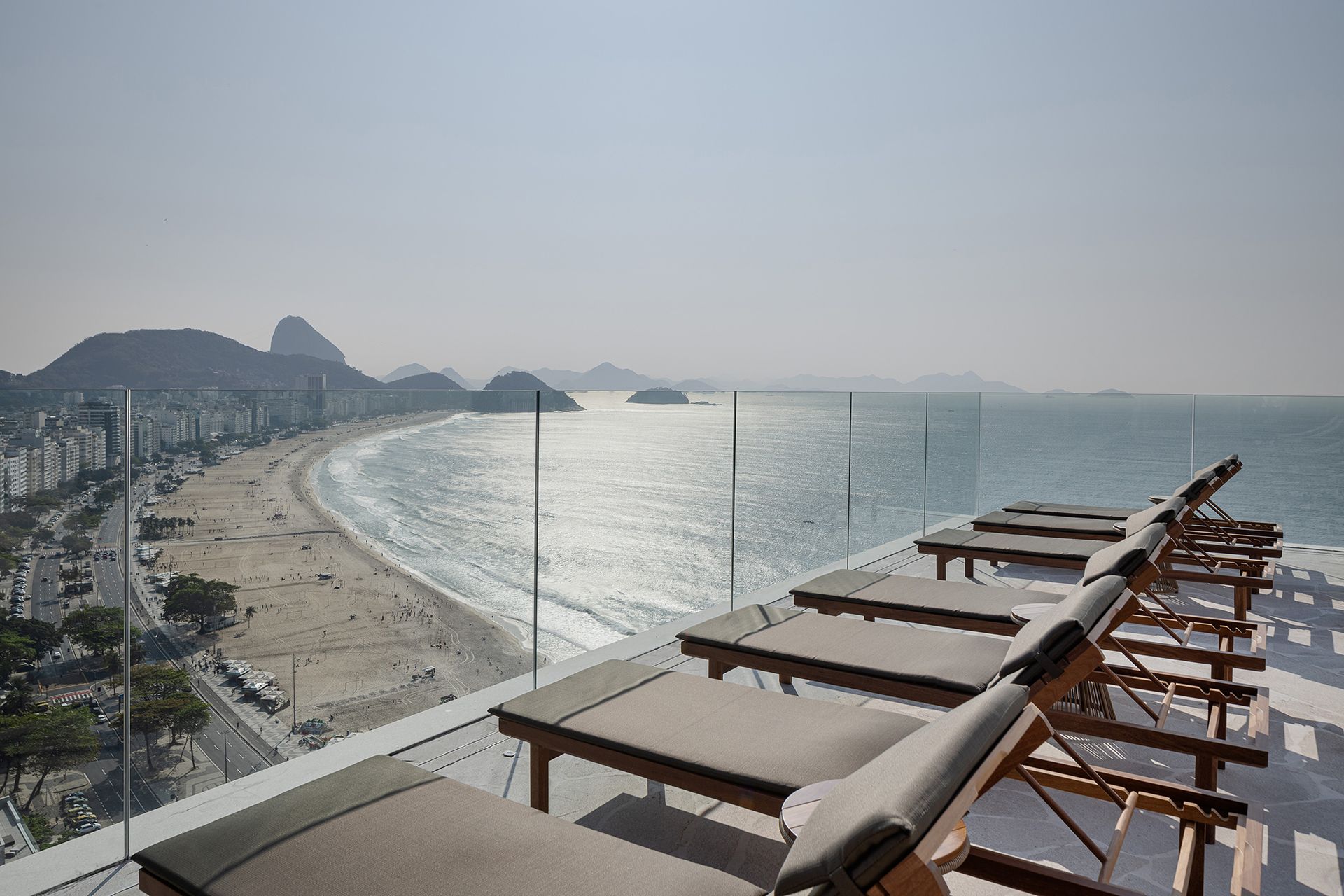 Terraço na cobertura com espreguiçadeiras com vista para uma praia ensolarada, o oceano e a cidade.