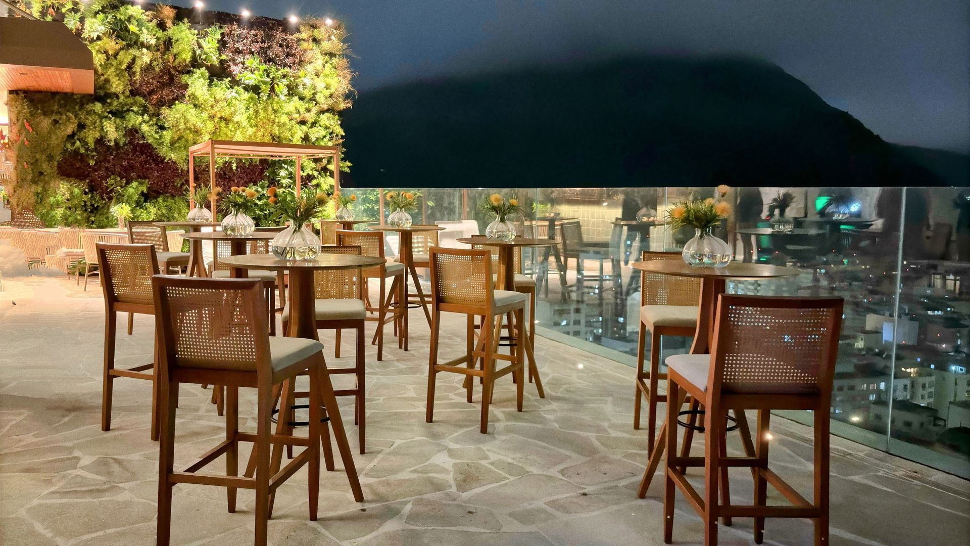 Bar na cobertura à noite com bancos de madeira, mesas e uma parede verde. Luzes da cidade e montanhas ao fundo. Othon