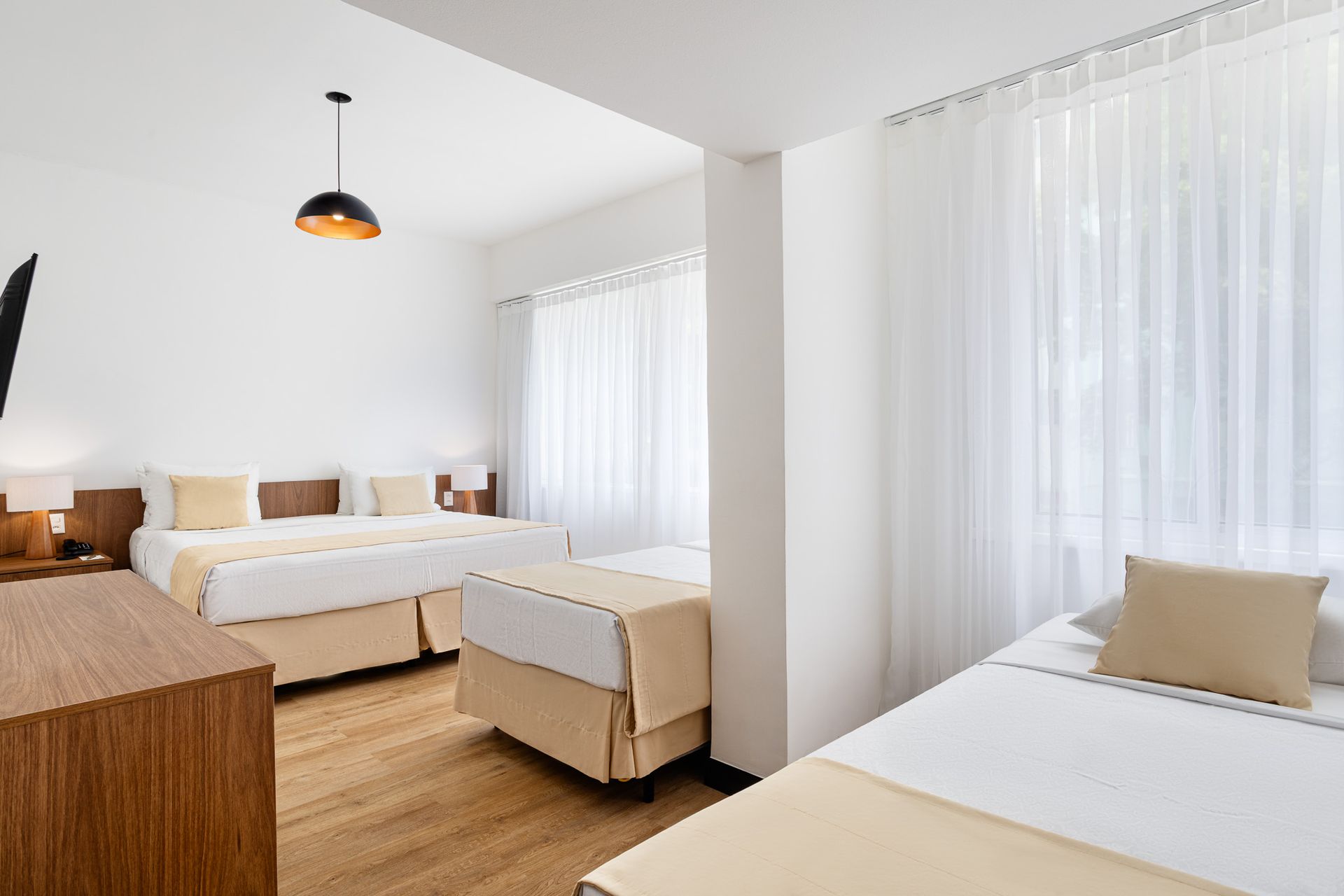 Quarto de hotel com três camas, móveis de madeira e cortinas brancas.