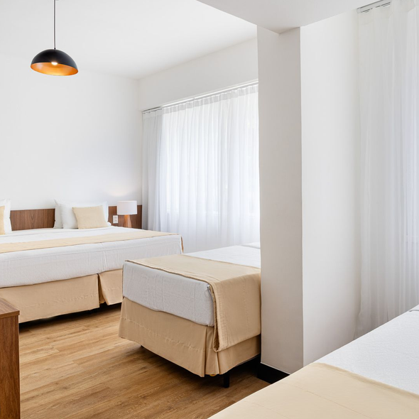 Quarto de hotel luminoso com várias camas e cortinas brancas.