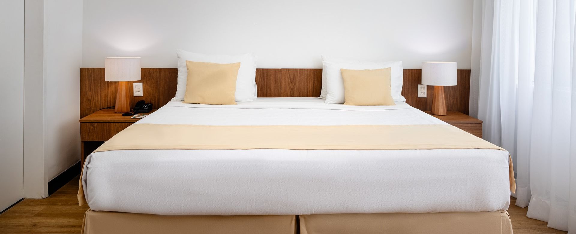 Uma cama de hotel bem arrumada com dois travesseiros, luminárias e uma parede branca.