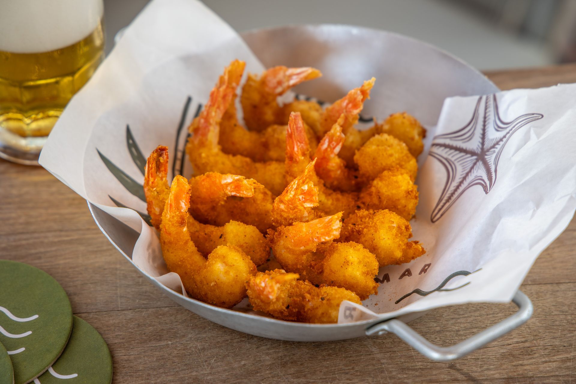 Camarões fritos e bolinhos de milho em um prato de metal, com uma cerveja ao fundo.