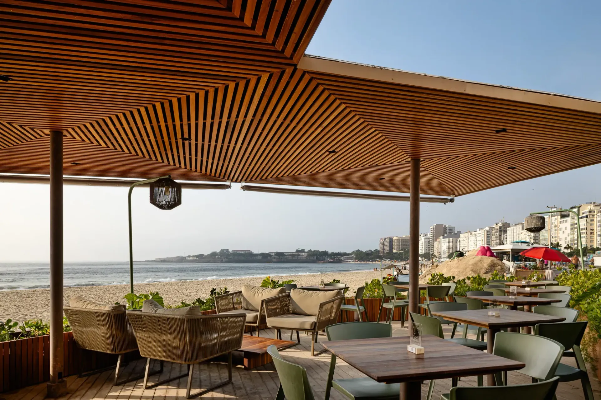 Restaurante com vista para a praia, cobertura de madeira, mesas, cadeiras e praia ao fundo.