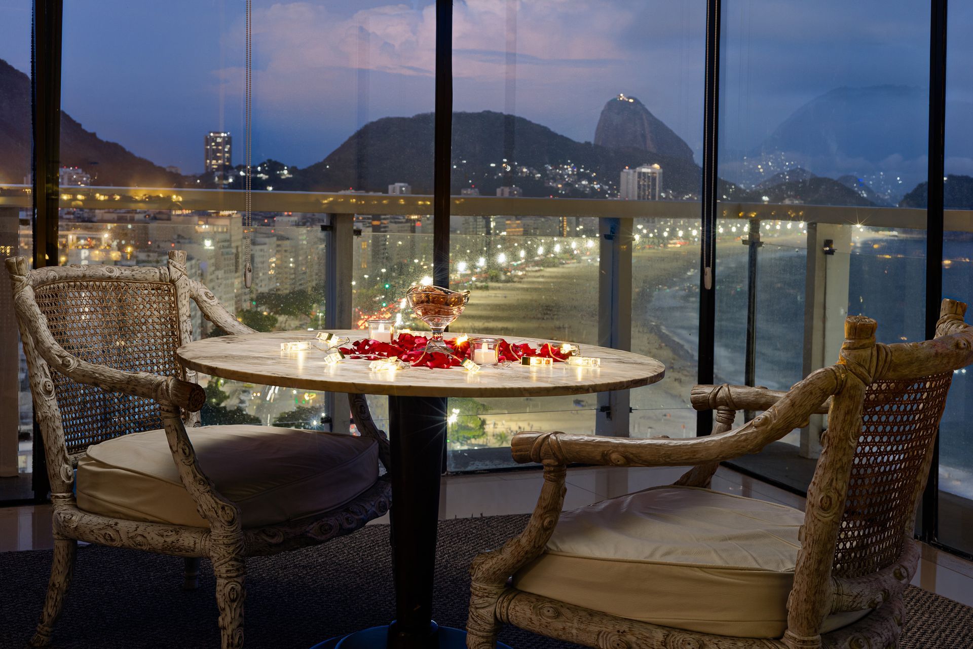 Mesa posta em um ambiente romântico com vista para a praia ao entardecer; rosas, velas, luzes da cidade.