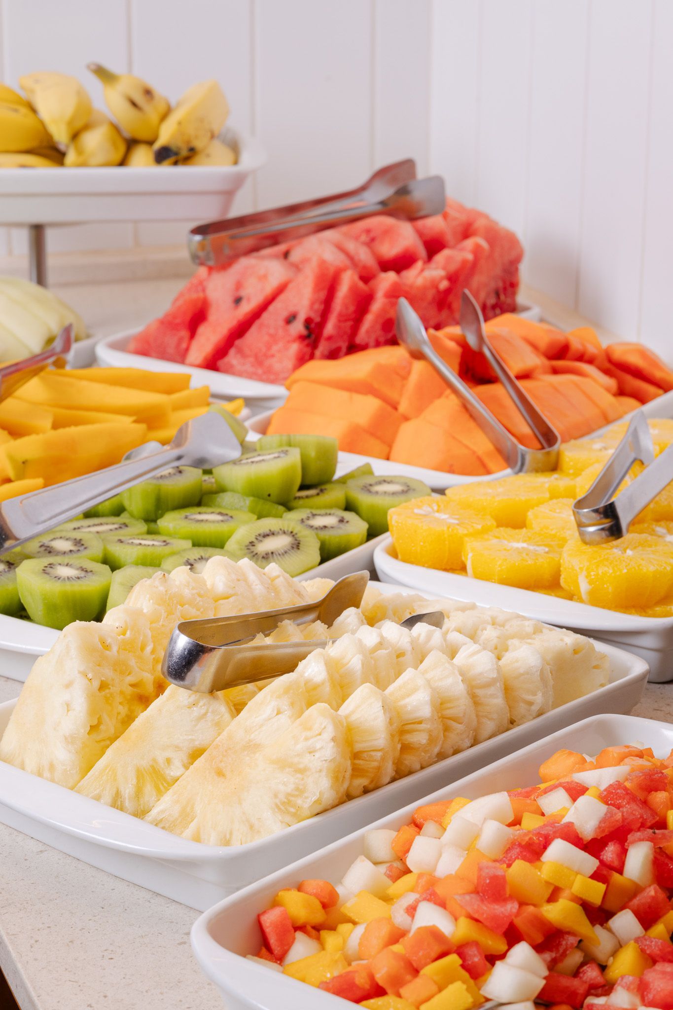 Buffet de frutas: bananas, melancia, mamão, kiwi, abacaxi e frutas picadas em bandejas brancas.