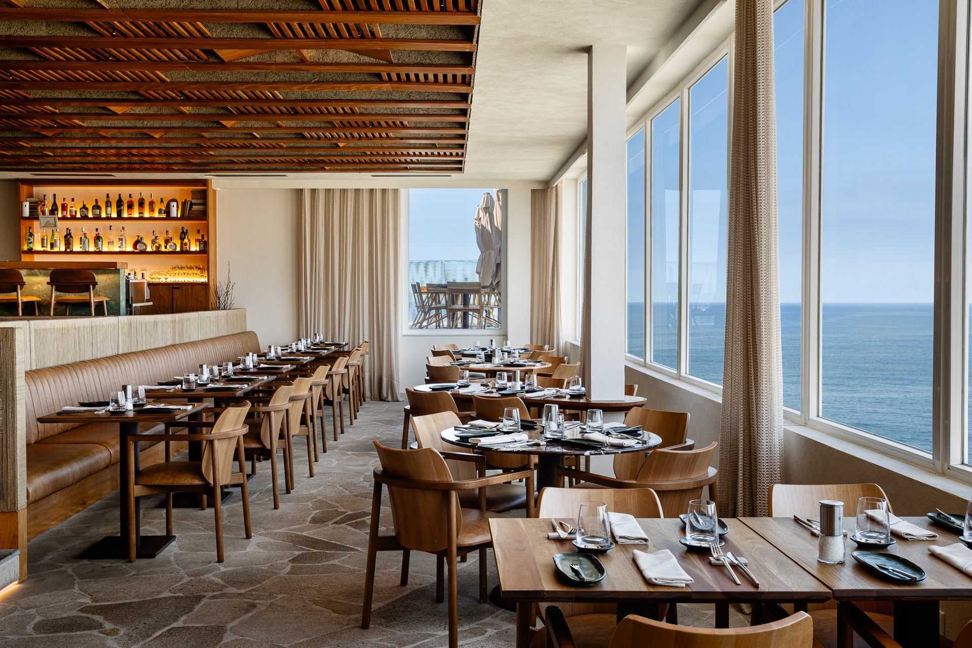Interior do restaurante com vista para o oceano; mesas e cadeiras de madeira, bar com garrafas de bebidas alcoólicas, janelas amplas.