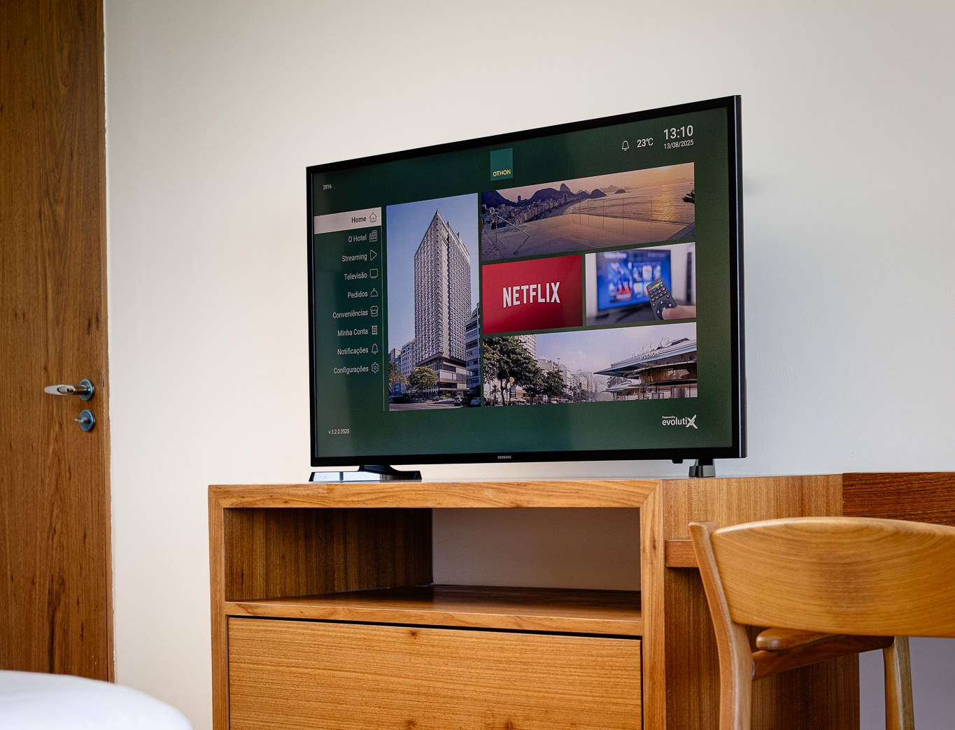 Uma televisão sobre um móvel de madeira exibe a tela inicial de um serviço de streaming. Uma cadeira e uma porta aparecem ao fundo.