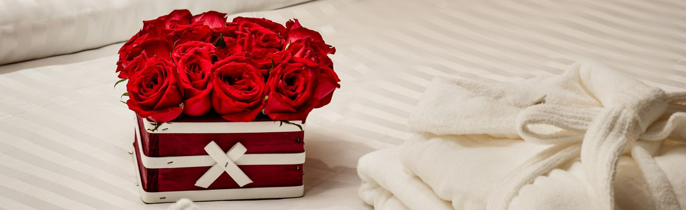 Rosas vermelhas em uma pequena caixa, colocadas sobre uma cama branca com uma toalha enrolada e amarrada com uma fita.