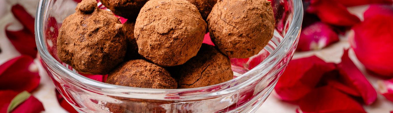 Trufas de chocolate em uma tigela de vidro, rodeadas por pétalas de rosa vermelha.