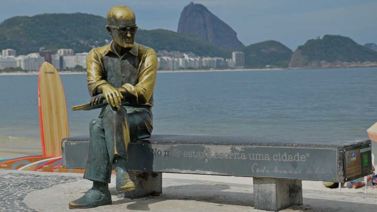 Estátua de um homem sentado em um banco, olhando para a Praia de Copacabana, Rio de Janeiro; Pão de Açúcar ao fundo.