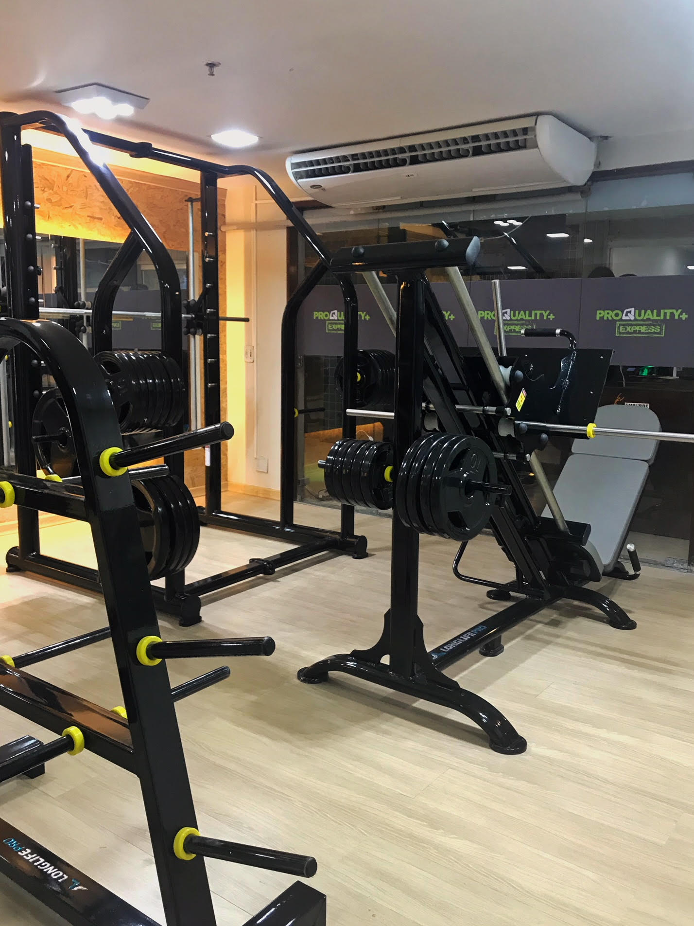 Interior de academia com equipamentos de musculação. Suportes de peso pretos e uma máquina de leg press estão dispostos em um piso de madeira.