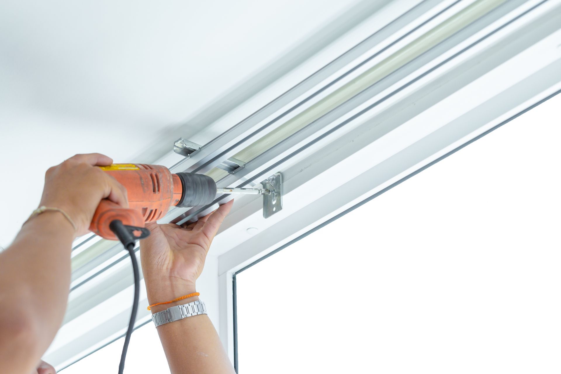 Person using power drill to install curtain rod bracket on a white ceiling above window.