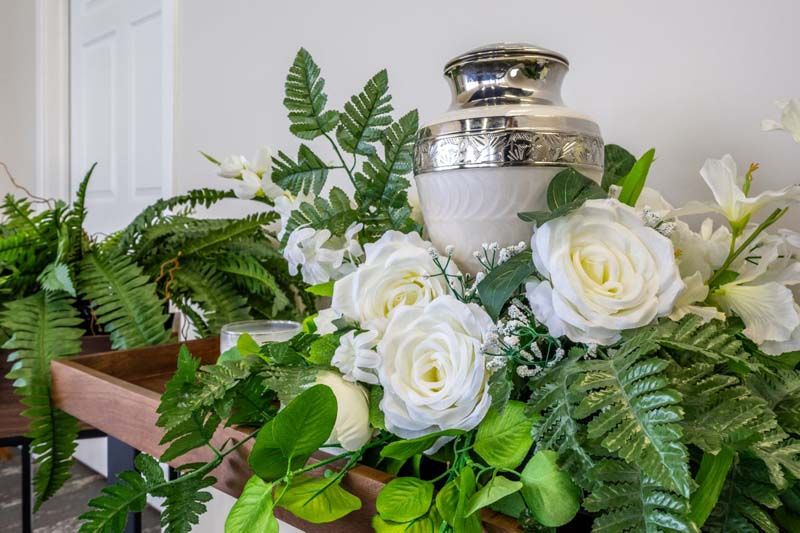 A cremation urn surrounded by white flowers and ferns on a table.