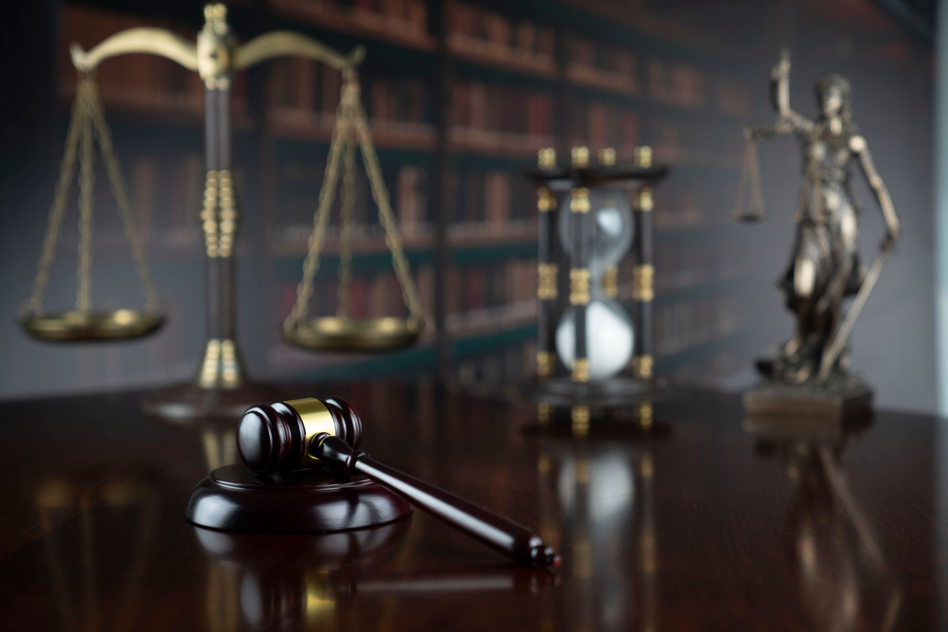 Gavel on table with scales of justice, hourglass, and Lady Justice statue in a library setting.
