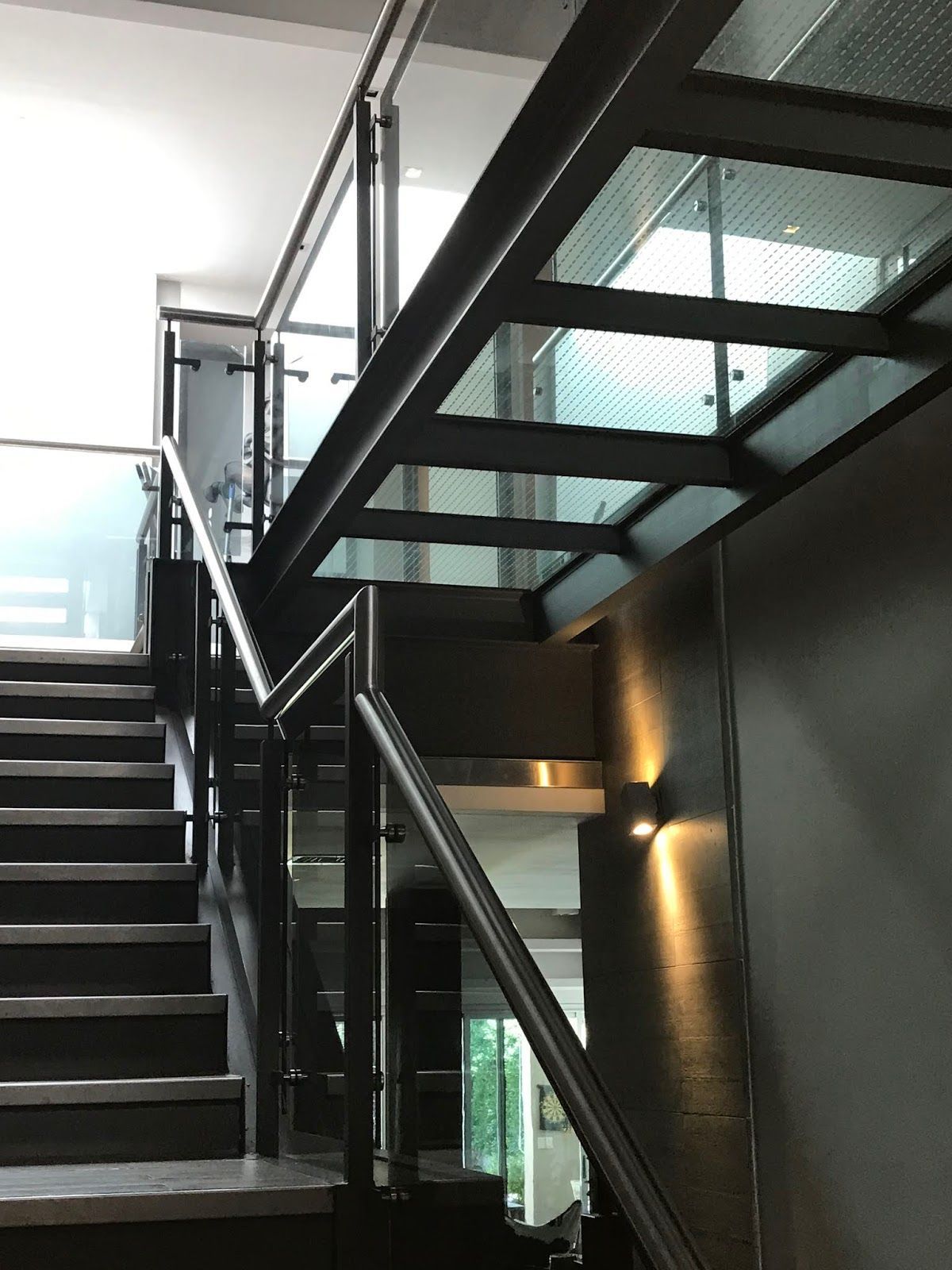 Escalera moderna con estructura de metal oscuro, barandillas de cristal y una lámpara de pared, vista desde abajo.