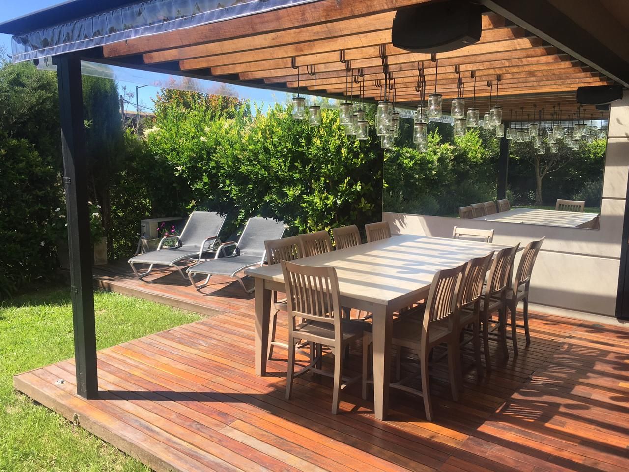 Una terraza de madera con una mesa de comedor y sillas bajo una pérgola, junto a dos tumbonas al lado de un seto verde.