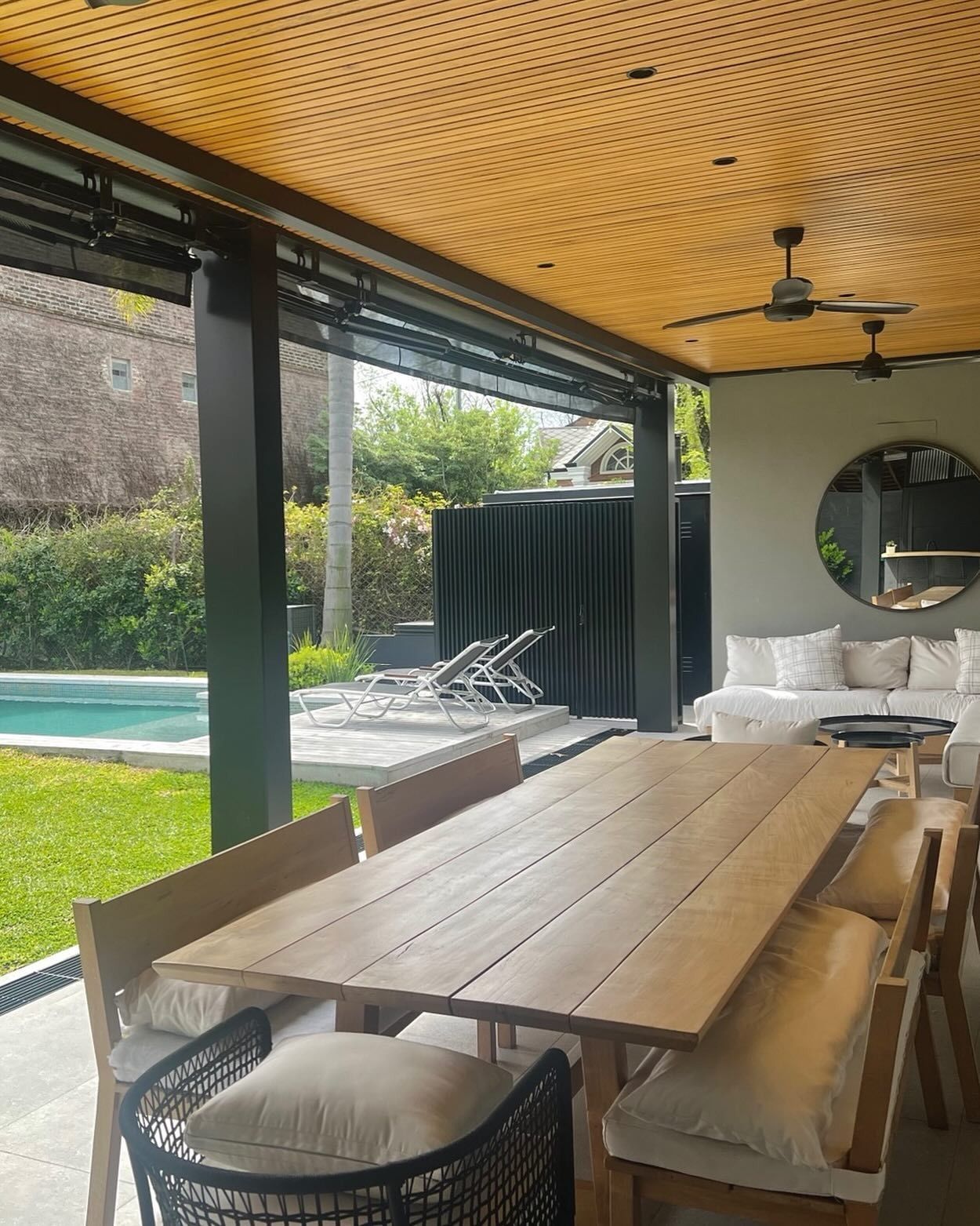Una mesa de comedor de madera y bancos en un patio cubierto, con una piscina al fondo y un techo con un estampado amarillo.