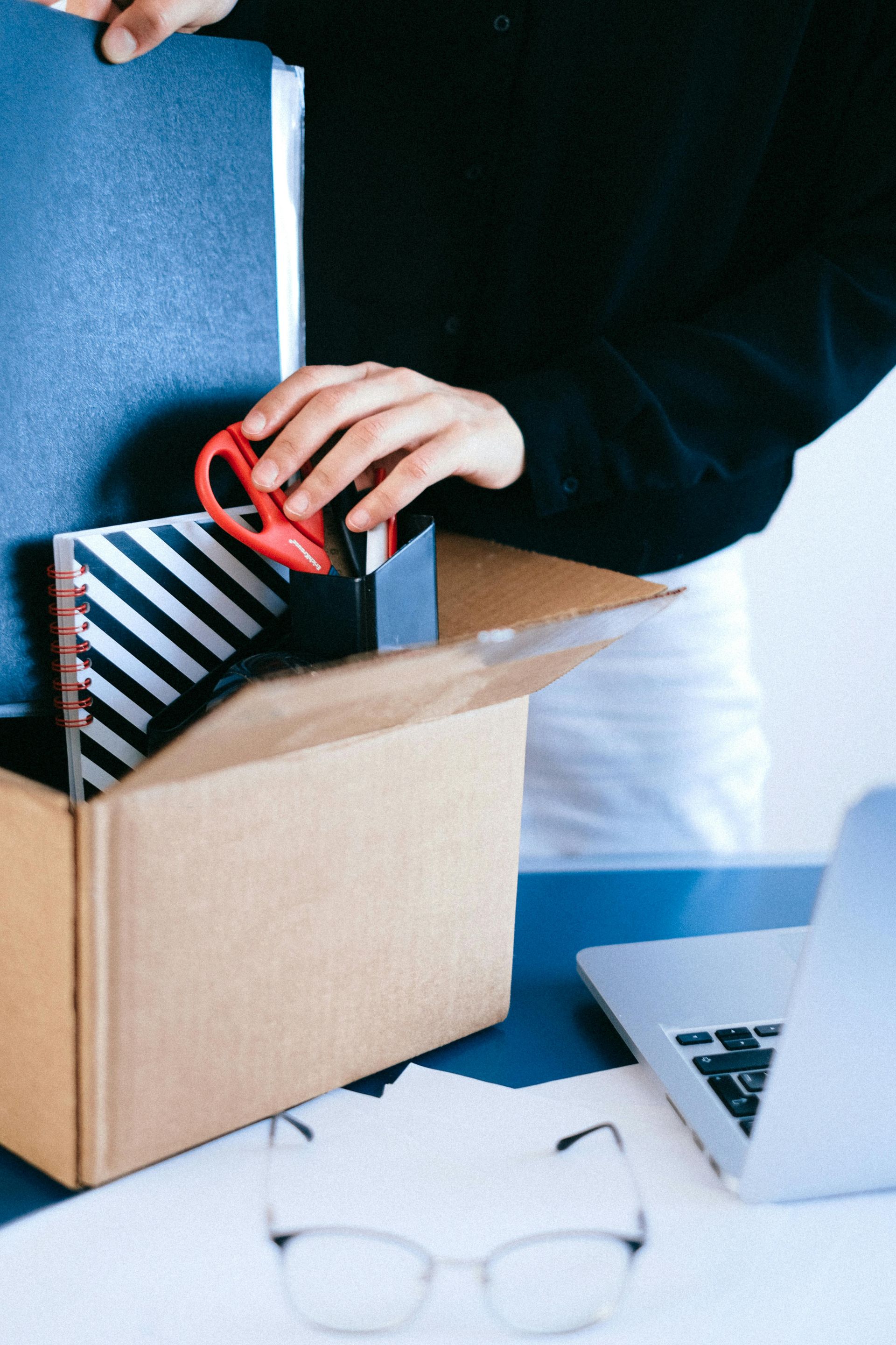 Person packing a cardboard box with books, a red object, and a notebook. A laptop and glasses are on the desk.