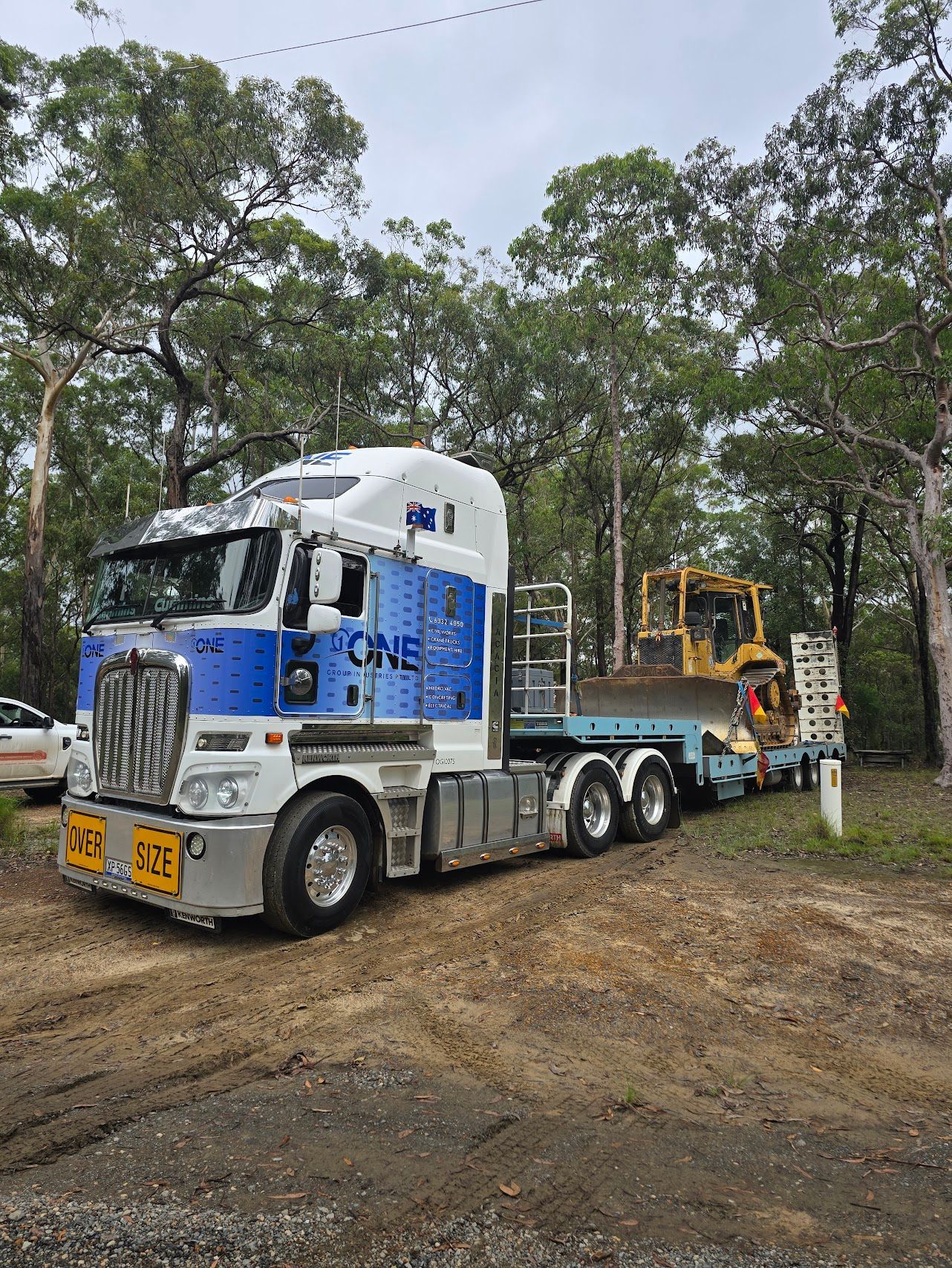 A large truck with a construction vehicle on a trailer parked in a wooded area — One Group Industries Pty Ltd in Bathurst, NSW