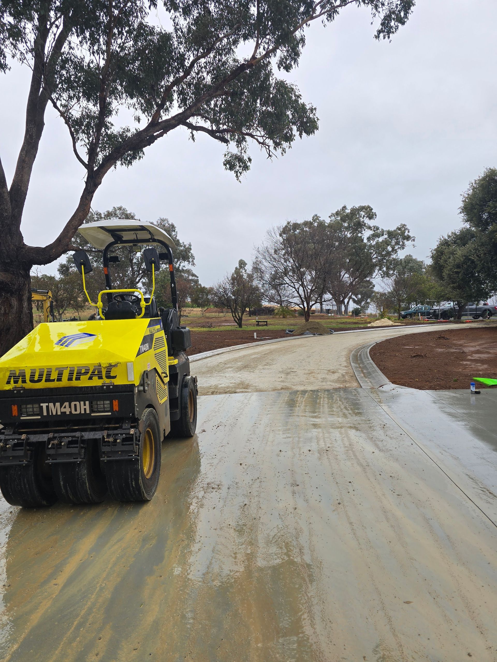 Yellow roller compacting wet, grey gravel on a winding road — One Group Industries Pty Ltd in Bathurst, NSW