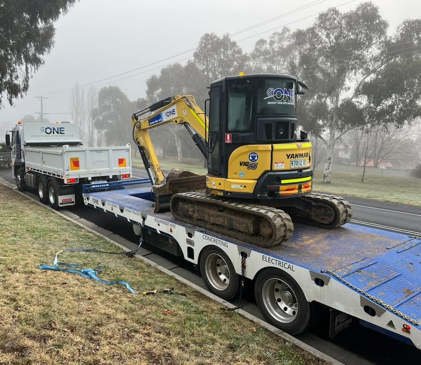 A yellow excavator on a flatbed trailer attached to a truck on a foggy road — One Group Industries Pty Ltd in Bathurst, NSW