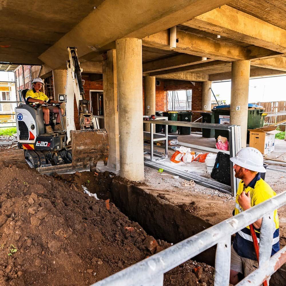 A Man is Kneeling in Front of a Mini Excavator at a Construction Site — One Group Industries Pty Ltd in Bathurst, NSW