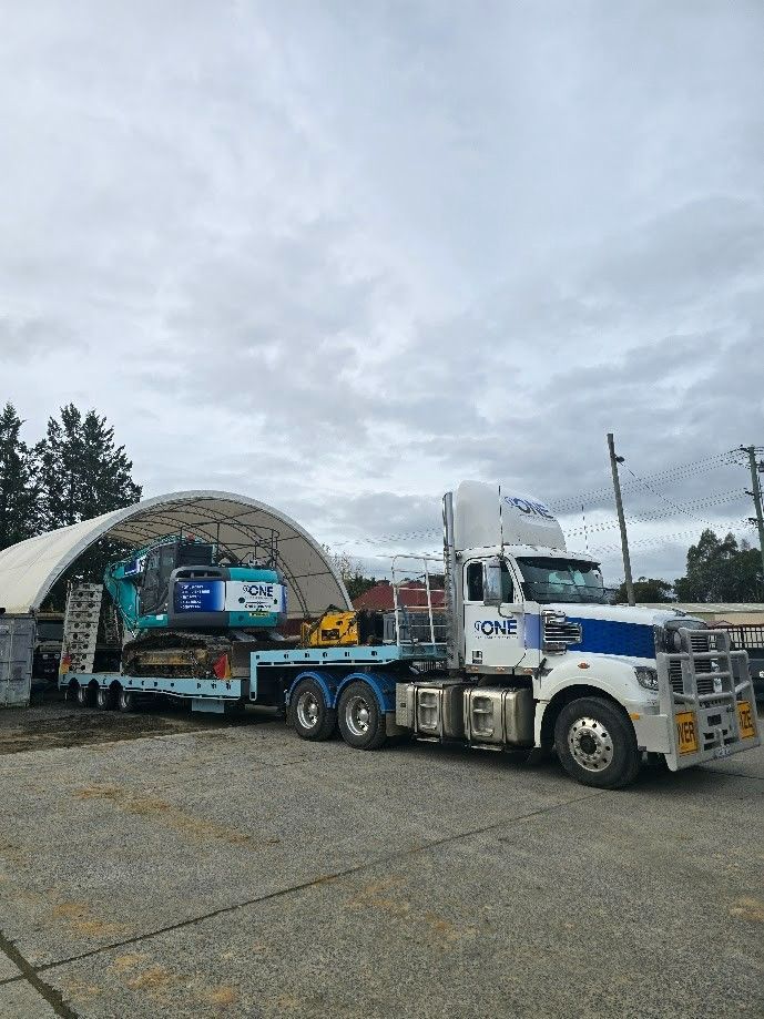 A Flatbed Truck Hauling an Excavator Under an Arched Shelter — One Group Industries Pty Ltd in Bathurst, NSW