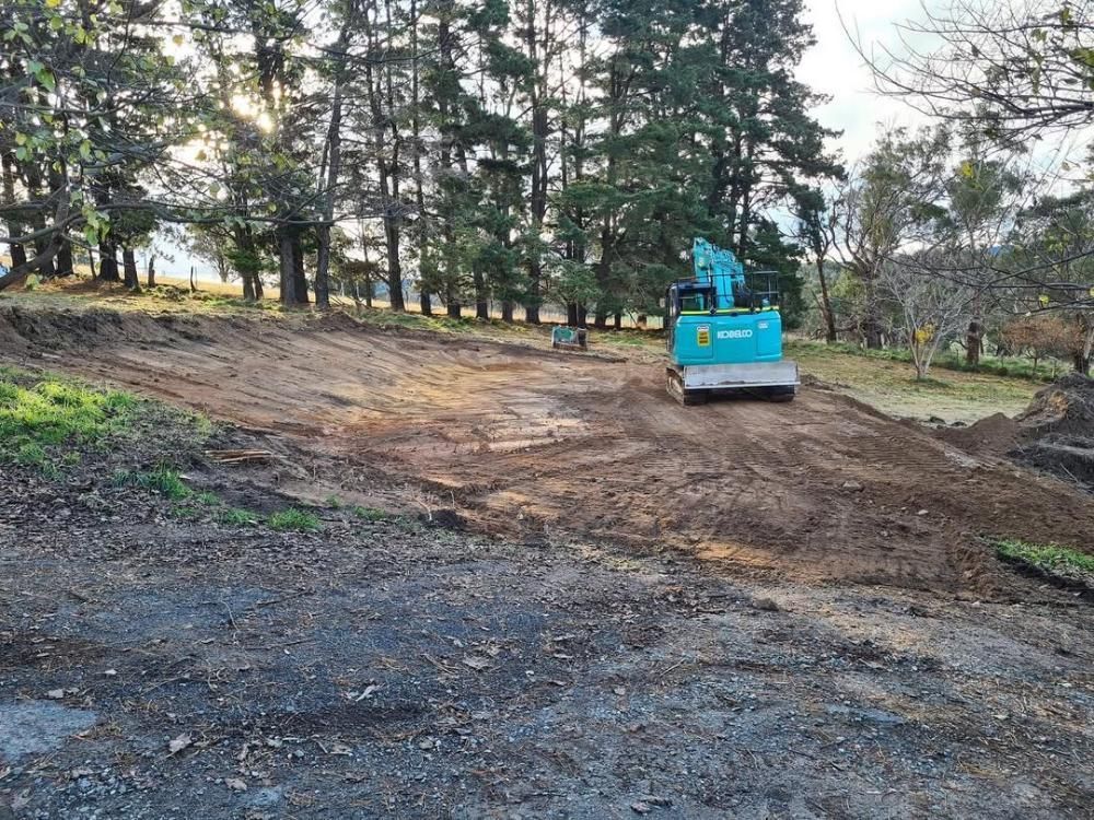 A Blue Excavator is Driving Down a Dirt Road Next to a Forest — One Group Industries Pty Ltd in Bathurst, NSW