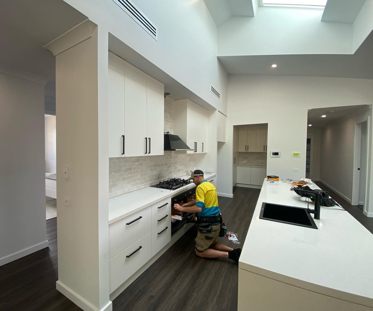 Person installing a gas cooktop in a white kitchen with dark cabinets and a skylight — One Group Industries Pty Ltd in Bathurst, NSW