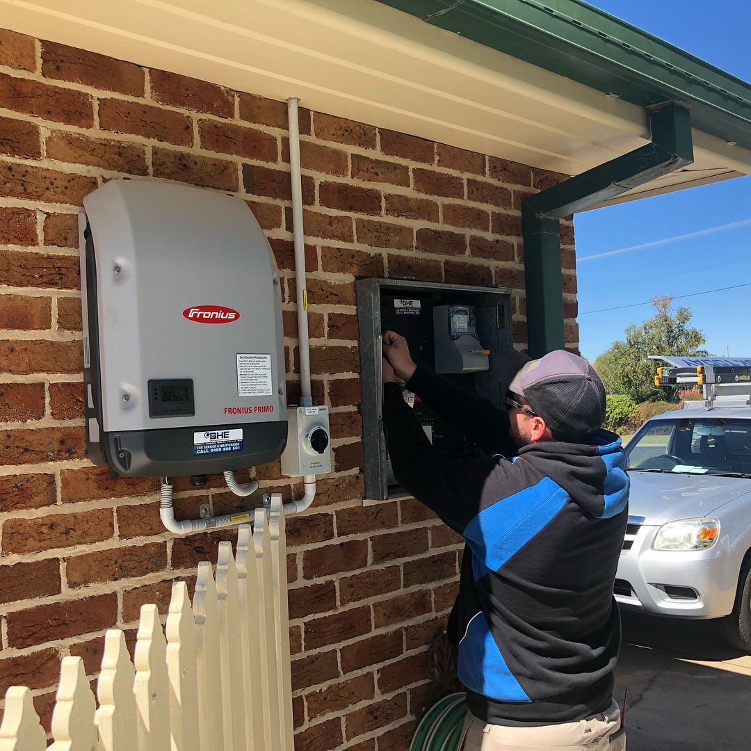 Man installing electrical equipment on a brick wall next to a gray solar panel inverter — One Group Industries Pty Ltd in Bathurst, NSW