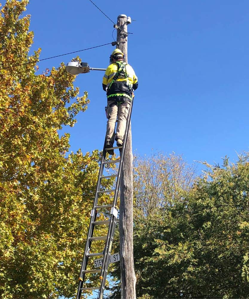 Lineman in yellow safety vest and helmet on a ladder on a utility pole, working outside on a sunny day — One Group Industries Pty Ltd in Bathurst, NSW
