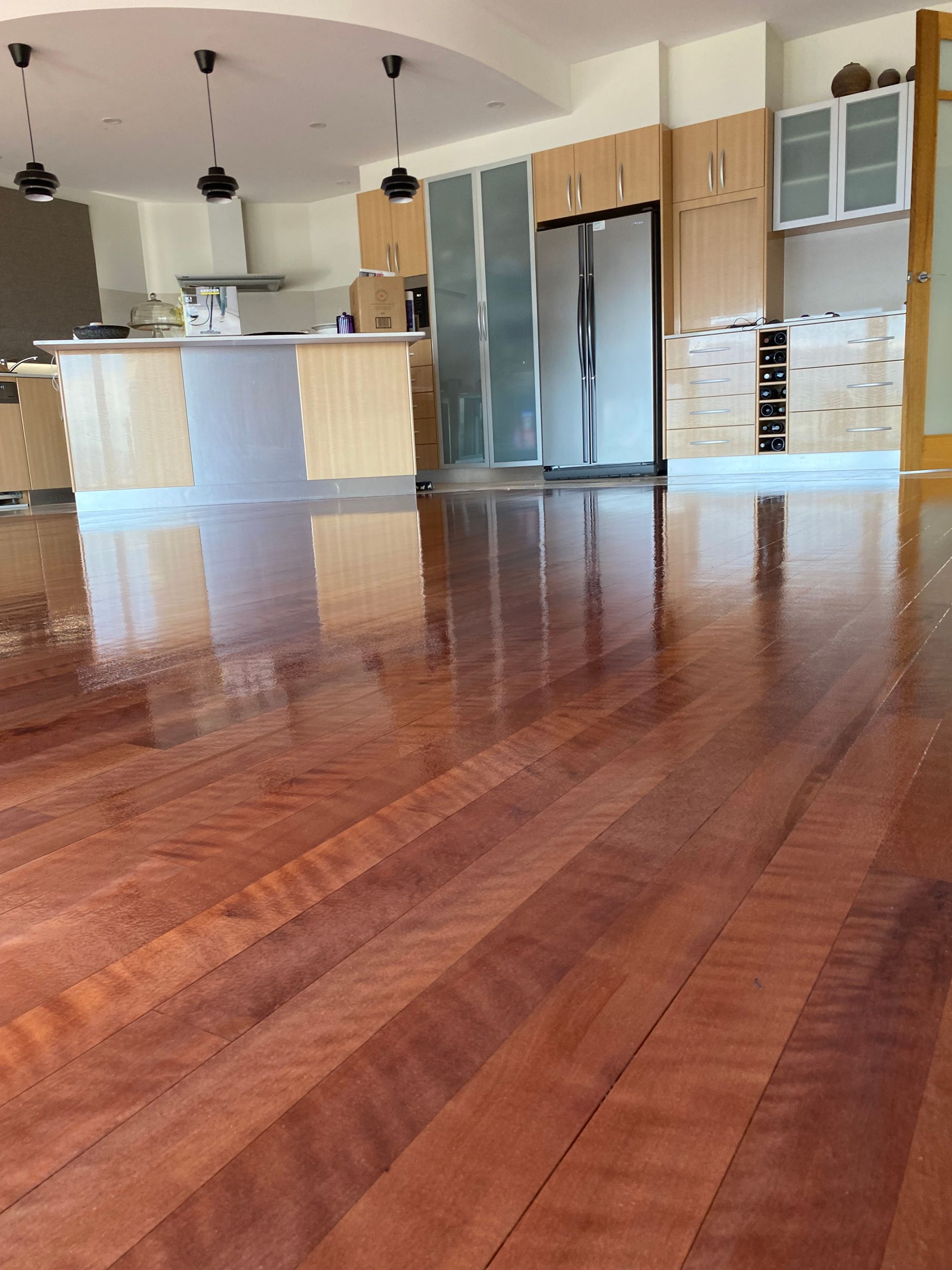 Empty kitchen with glossy reddish wood floors, island, stainless steel fridge, and hanging pendant lights — Finer Floors In Cambridge, TAS