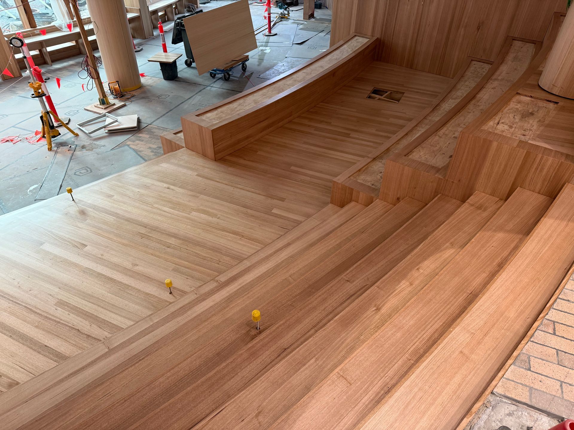 A construction site showing tiered wooden platforms and stairs under assembly, with light wood tones and a concrete floor.