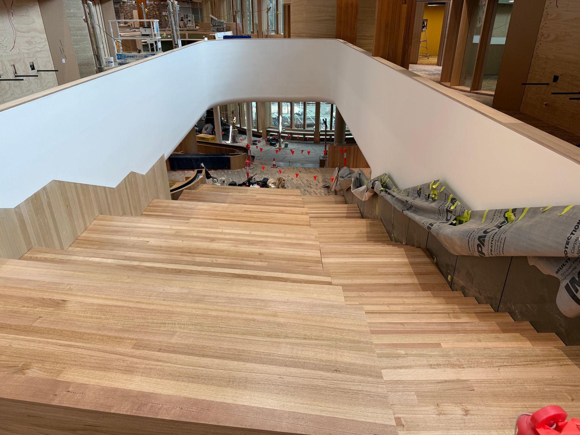 Wide wooden staircase in a modern building atrium, with a curved white wall and lower seating area below. — Finer Floors In Cambridge, TAS
