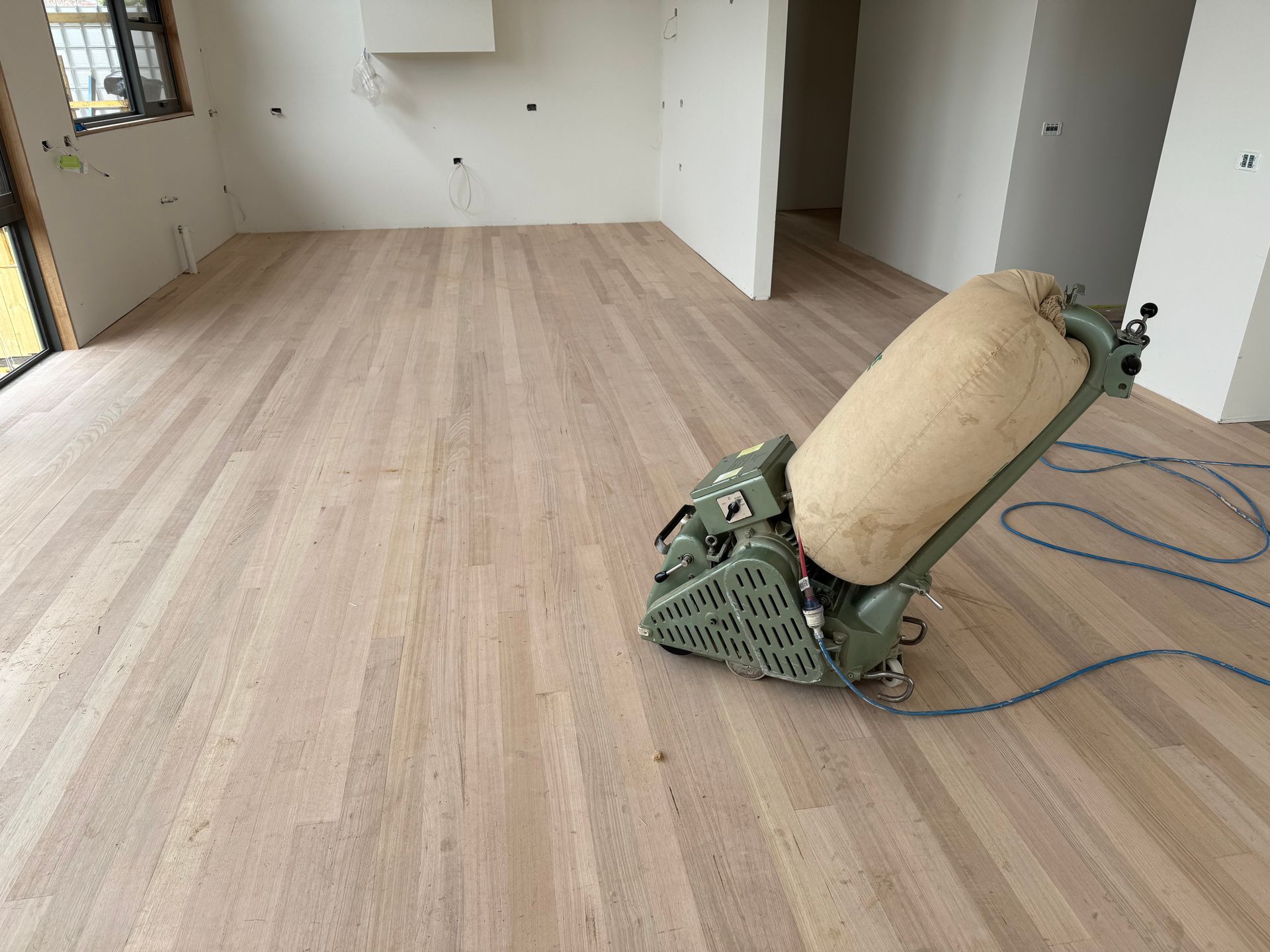 A floor sanding machine sits on a newly sanded light-colored hardwood floor in an unfinished room.