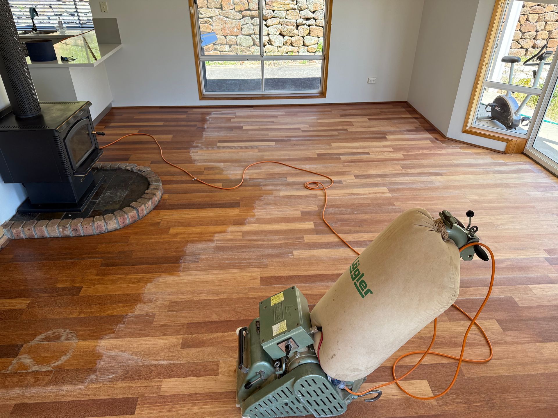 Floor sander in a room with a partially refinished hardwood floor and a fireplace. — Finer Floors In Cambridge, TAS