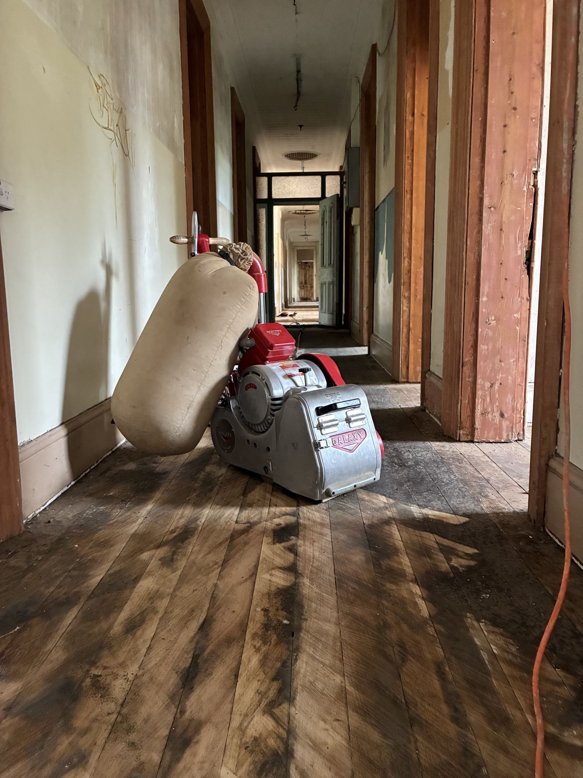 Narrow hallway with damaged wooden floors and scattered debris, including a leaning cushion and a vacuum cleaner — Finer Floors In Cambridge, TAS