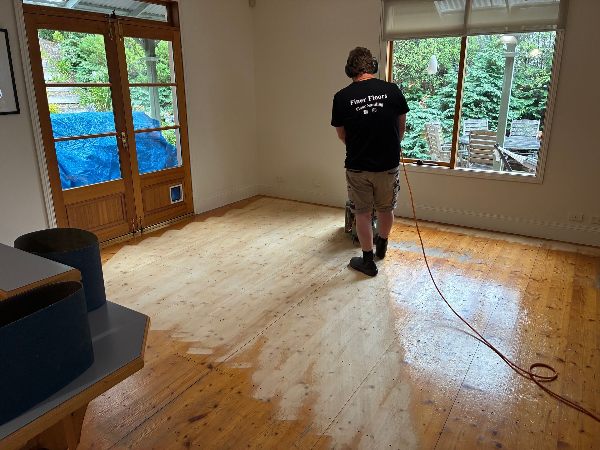Person sanding a wooden floor in a bright room with large windows and French doors. — Finer Floors In Cambridge, TAS