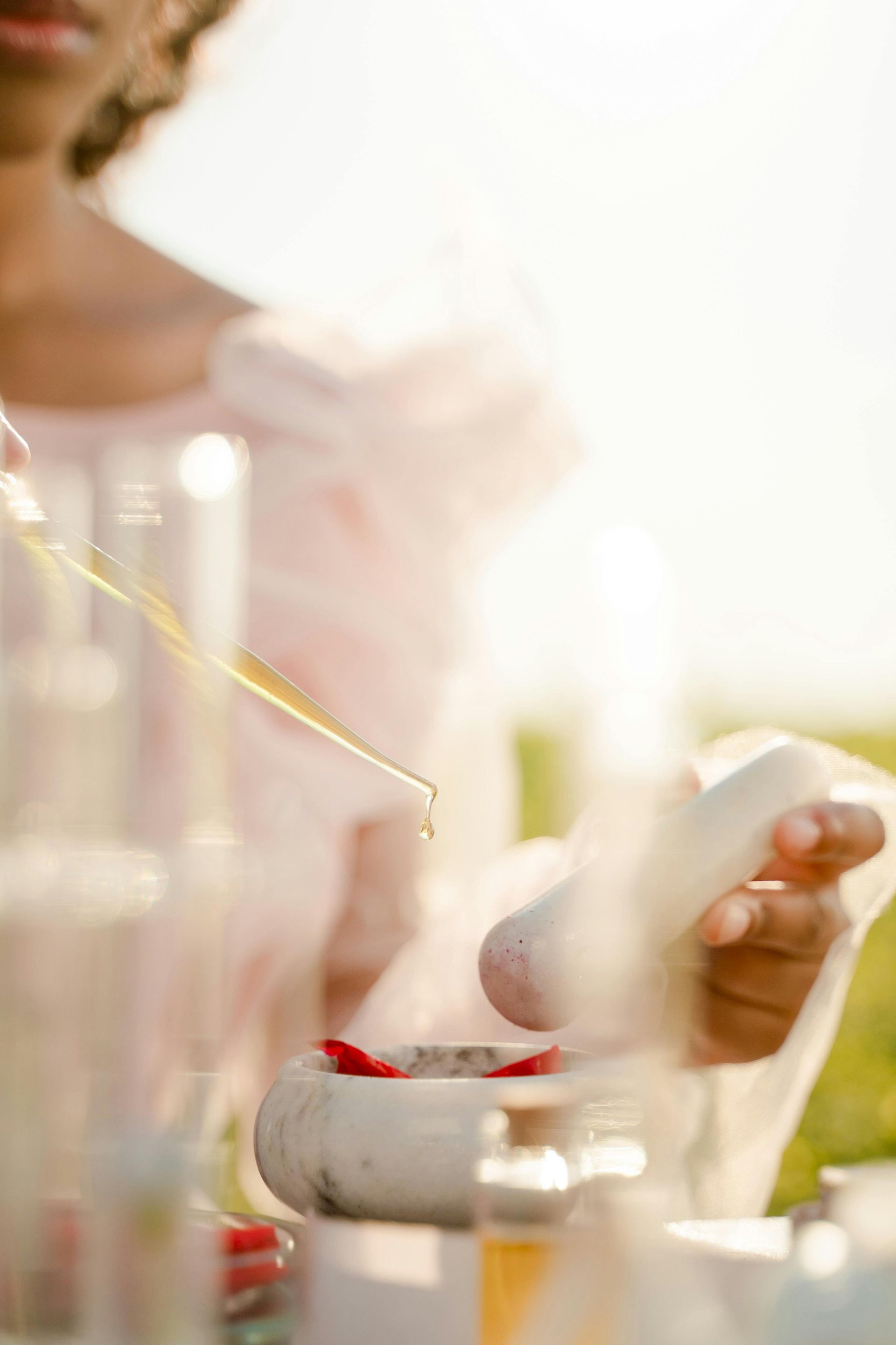 Person holding a dropper over a mortar and pestle, outdoors, light pink clothing.
