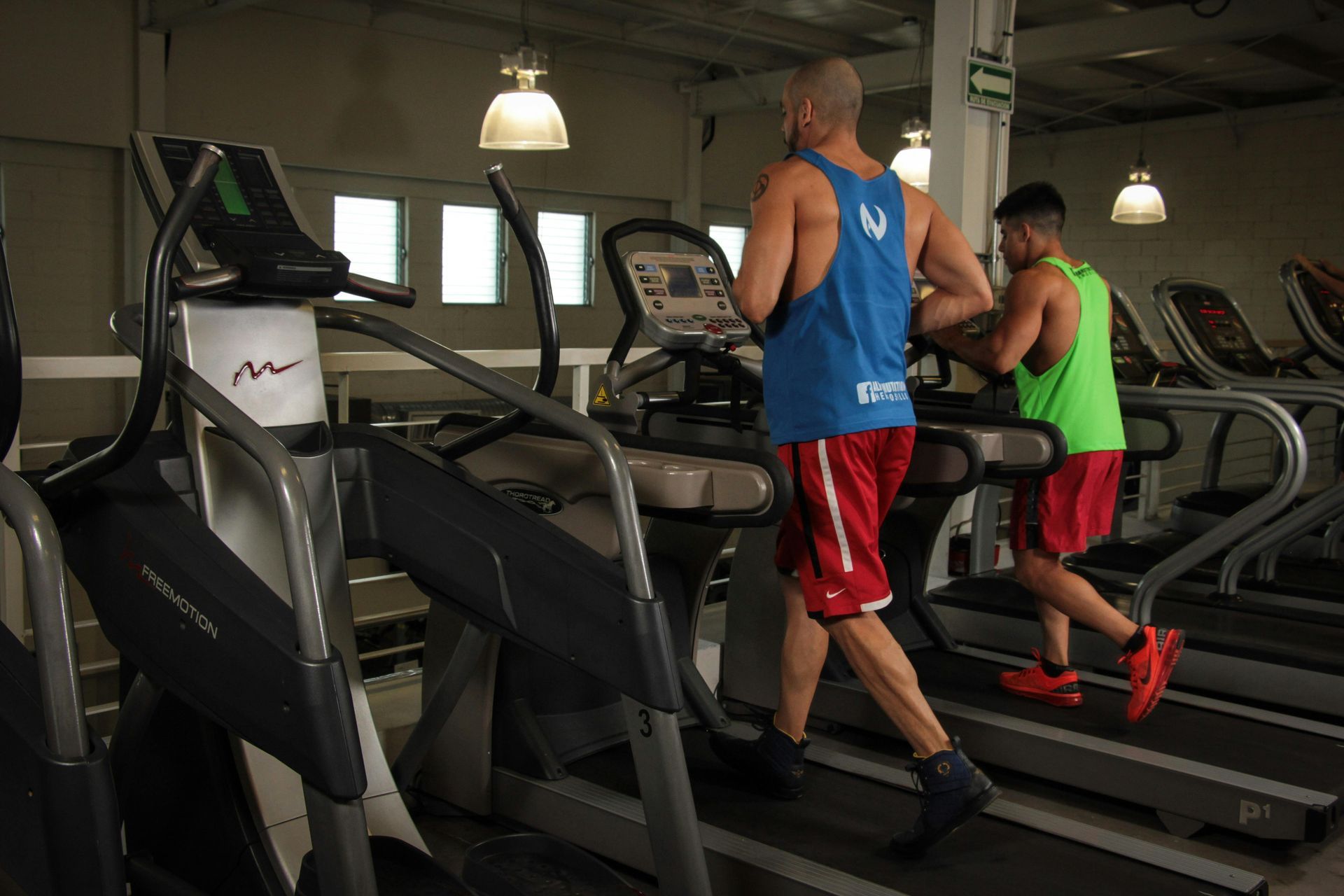 Two people running on treadmills in a gym. One wears blue, the other green; both wear red shorts.
