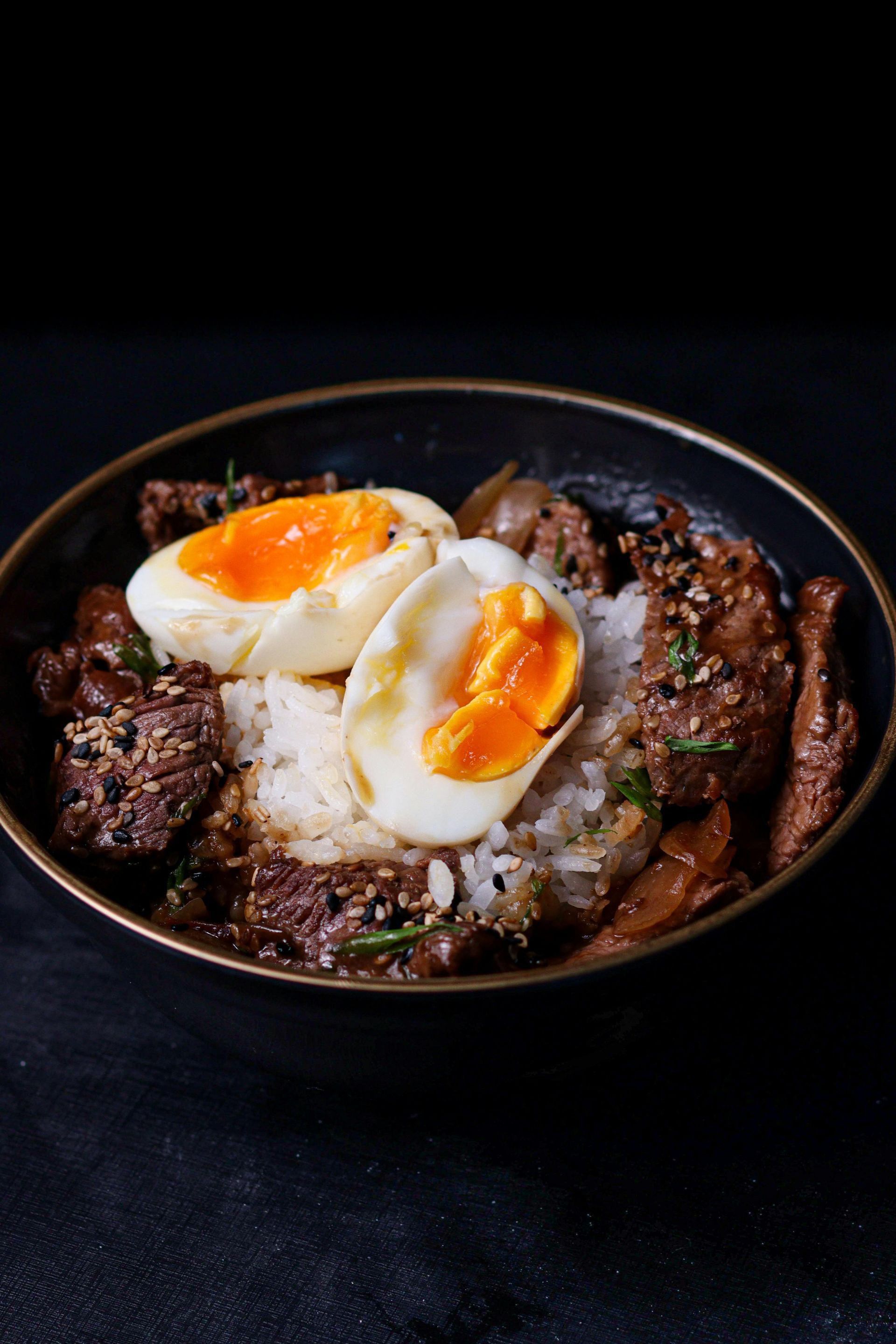 Bowl of rice topped with braised beef, soft-boiled egg halves with orange yolks, and sesame seeds.